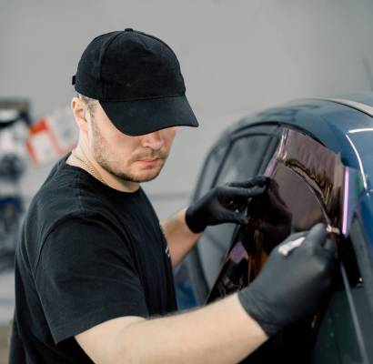 Closeup image of a handsome car mechanic worker, wearing black uniform, attaching tinting foil to car window in specialized service station.